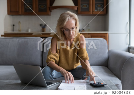 Serious older woman calculates household finances, paying bills on laptop Serious older woman calculates household finances, paying bills on laptop 92004338