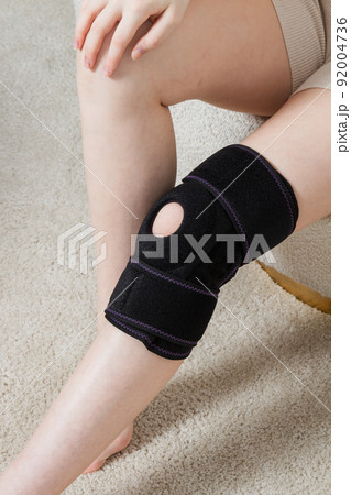 Black knee support brace on a woman leg. girl in an orthosis in the interior of the house. Orthopedic anatomic braces for knee fixation, injuries and pain. Knee Joint Bandage Sleeve. Elastic Sports. 92004736