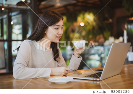 Asian beautiful working woman doing her work and drink in the coffee shop or cafe. Cheerful online blogger writing on her laptop or notebook computer and drinking a coffee close up. Modern lifestyle. Asian beautiful working woman doing her work and drink in the coffee shop or cafe. Cheerful online blogger writing on her laptop or notebook computer and drinking a coffee close up. Modern lifestyle. 92005286