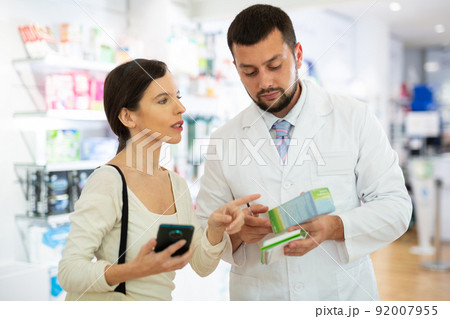 Female shopper shows name of drug on smartphone screen. Pharmacist helps to find medicine in pharmacy Female shopper shows name of drug on smartphone screen. Pharmacist helps to find medicine in pharmacy 92007955