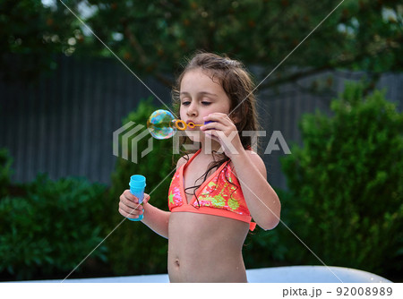 Caucasian funny little girl, blows soap balls in the inflatable water pool on summer vacation. 92008989