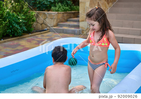 Happy kids splash and play ball in the pool on hot summer day. Summer activities for kids. Water fun day in a backyard. Happy kids splash and play ball in the pool on hot summer day. Summer activities for kids. Water fun day in a backyard. 92009026