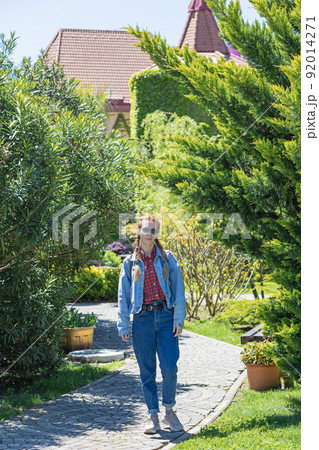 A smiling woman in a denim outfit walks along a paved park path passing between lawns planted with dense trees and bushes on a sunny spring day 92014271