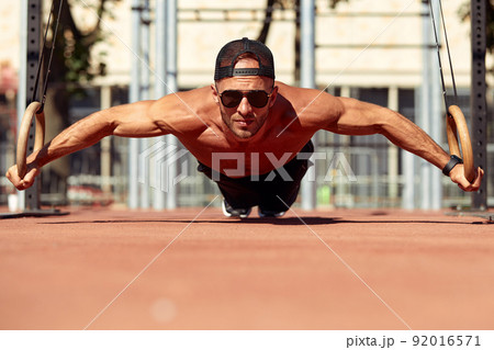 Handsome man on sport ground. The sportsman the guy, carries out difficult exercise, sports gymnastics. Muscular man doing workout with rings. The concept of a modern young man Handsome man on sport ground. The sportsman the guy, carries out difficult exercise, sports gymnastics. Muscular man doing workout with rings. The concept of a modern young man 92016571