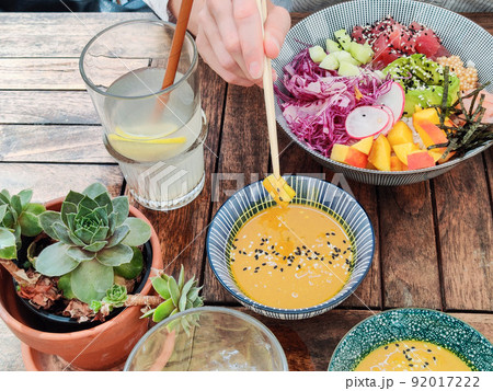 Woman eating tasty colorful healthy natural organic vegetarian Hawaiian poke bowl using asian chopsticks on rustic wooden table. Healthy natural organic eating concept Woman eating tasty colorful healthy natural organic vegetarian Hawaiian poke bowl using asian chopsticks on rustic wooden table. Healthy natural organic eating concept 92017222