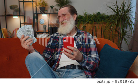 Smiling happy senior old grandfather counting money cash use smartphone, income, saves, lottery win 92019735