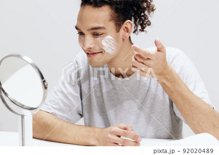 handsome guy applying cream on the face and looking in the mirror cosmetology cropped view 92020489