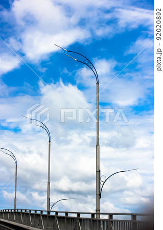 Modern LED lamp posts in a row against a blue sky with white clouds vertical wallpaper. Road lighting, electric streetlights abstract background. City illumination in daylight. Light off. Industry. Modern LED lamp posts in a row against a blue sky with white clouds vertical wallpaper. Road lighting, electric streetlights abstract background. City illumination in daylight. Light off. Industry. 92020892