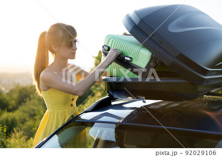 Young woman taking green suitcase out from car roof rack. Travel and vacations concept. Young woman taking green suitcase out from car roof rack. Travel and vacations concept. 92029106