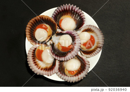 A white plate of big raw fresh scallops with orange caviar and shells on a dark background.  92036940