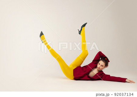Portrait of young stylish girl in yellow tights, red jacket and heel shoes posing on floor isolated over grey studio background. 92037134