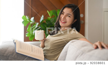 Cheerful asian woman in casual clothes holding cup of tea in her hand, relaxing on couch at home 92038601