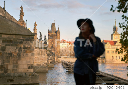 Prague Silhouette. Stylish beautiful young woman wearing black hat with Charles Bridge on background. Elegant retro lady fine art portrait. Focus on background. 92042401