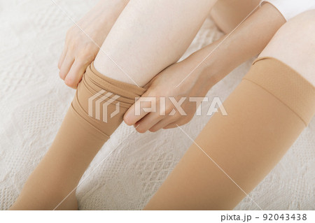Beautiful long female legs in stockings. Girl putting on stockings at home in a white room. Beige knee socks. Varicose veins prevention. Woman body in underwear. 92043438