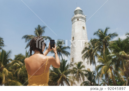 Woman traveler in dress in front of famous landmark of Sri Lanka country taking photo on smartphone of Dondra lighthouse Woman traveler in dress in front of famous landmark of Sri Lanka country taking photo on smartphone of Dondra lighthouse 92045897