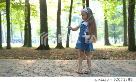 Happy little child girl blowing soap bubbles outside in green park. Outdoor summer activities for children concept. Happy little child girl blowing soap bubbles outside in green park. Outdoor summer activities for children concept. 92051599
