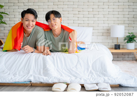Couple gay or lgbt man use rainbow flag to cover and lie on white bed also look at camera with smile Couple gay or lgbt man use rainbow flag to cover and lie on white bed also look at camera with smile 92052087