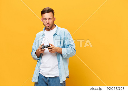 Serious concentrated tanned handsome man in blue basic t-shirt play new game with joystick posing isolated on orange yellow studio background. Copy space Banner Mockup. People lifestyle concept 92053393