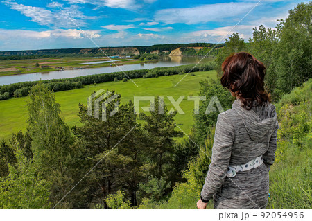 A girl standing on a hill, on a steep bank of a river in summer and looking into far away. 92054956