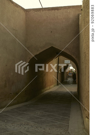A narrow street with adobe houses in the old city of Yazd, which barely fit people and a car 92069883