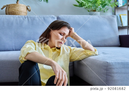 Sad upset middle-aged woman sitting on the floor near the sofa Sad upset middle-aged woman sitting on the floor near the sofa 92081682