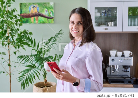 Young woman in kitchen with cup of coffee smartphone, looking at camera 92081741