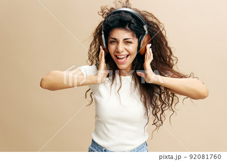 Music offer. Overjoyed Latin female with afro in wireless headphones enjoy playlist, sing and dance staying isolated near beige wall. Copy free space for design ad. Fashion IT Seasonal Sale concept Music offer. Overjoyed Latin female with afro in wireless headphones enjoy playlist, sing and dance staying isolated near beige wall. Copy free space for design ad. Fashion IT Seasonal Sale concept 92081760
