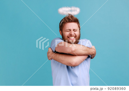 Portrait of bearded angelic man with holy nimbus standing smiling contentedly and hugging himself, self-love, support, keeps eyes closed. Indoor studio shot isolated on blue background. Portrait of bearded angelic man with holy nimbus standing smiling contentedly and hugging himself, self-love, support, keeps eyes closed. Indoor studio shot isolated on blue background. 92089360