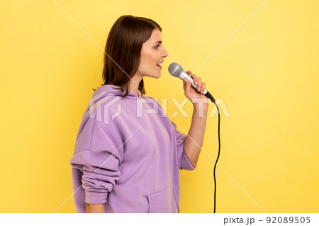 Side view of young beautiful woman reporter holding microphone, reporting and telling opinion, discussing important topics, wearing purple hoodie. Indoor studio shot isolated on yellow background. 92089505