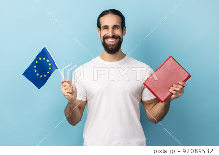 Portrait of satisfied smiling bearded man wearing white T-shirt holding book and europe flag, education abroad, looking at camera with toothy smile. Indoor studio shot isolated on blue background. 92089532