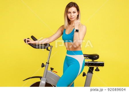 Portrait of blonde woman doing cardio exercises, standing and showing her smartwatch or fitness tracker, wearing blue sportswear. Indoor studio shot isolated on yellow background. Portrait of blonde woman doing cardio exercises, standing and showing her smartwatch or fitness tracker, wearing blue sportswear. Indoor studio shot isolated on yellow background. 92089604