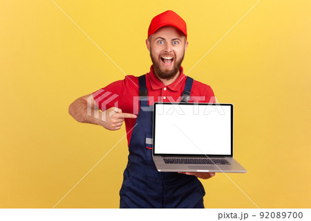 Portrait of excited happy worker man in uniform standing and pointing at laptop blank screen, advertisement area, looking smiling. Indoor studio shot isolated on yellow background. 92089700