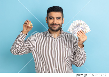 Happy businessman holding showing dollar bills and golden coin of crypto currency, transferring money into digital bitcoins, wearing striped shirt. Indoor studio shot isolated on blue background. 92089765