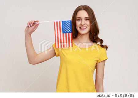 Smiling attractive teenager girl in yellow casual T-shirt holding usa flag looking at camera with toothy smile, human rights, freedom of speech. Indoor studio shot isolated on gray background. Smiling attractive teenager girl in yellow casual T-shirt holding usa flag looking at camera with toothy smile, human rights, freedom of speech. Indoor studio shot isolated on gray background. 92089788