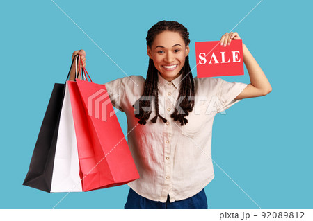 Attractive woman with black dreadlocks holding shopping bags and showing sale card, advertising low prices in the mall, wearing white shirt. Indoor studio shot isolated on blue background. Attractive woman with black dreadlocks holding shopping bags and showing sale card, advertising low prices in the mall, wearing white shirt. Indoor studio shot isolated on blue background. 92089812