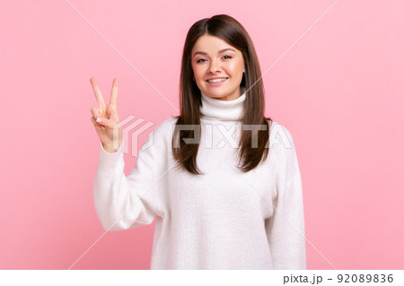 Positive dark haired young female smiling happily and showing v sign or peace gesture, rejoicing win, wearing white casual style sweater. Indoor studio shot isolated on pink background. 92089836