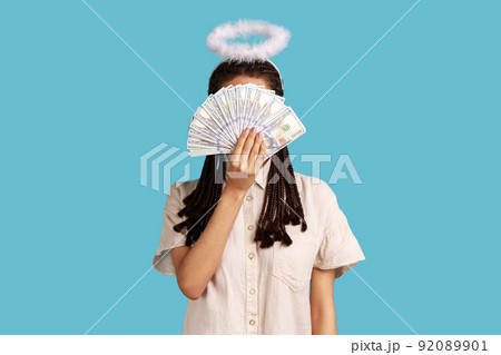 Angelic rich woman with black dreadlocks and nimbus on head hiding behind dollar banknotes, big profit earning cash, wearing white shirt. Indoor studio shot isolated on blue background. 92089901