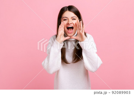 Portrait of young adult brunette female standing, holding arms near wide open mouth and screaming, wearing white casual style sweater. Indoor studio shot isolated on pink background. 92089903