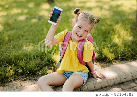 A charming little girl with a backpack is sitting on the sidewalk after school and showing her phone. Back to school, after school, school age A charming little girl with a backpack is sitting on the sidewalk after school and showing her phone. Back to school, after school, school age 92104383