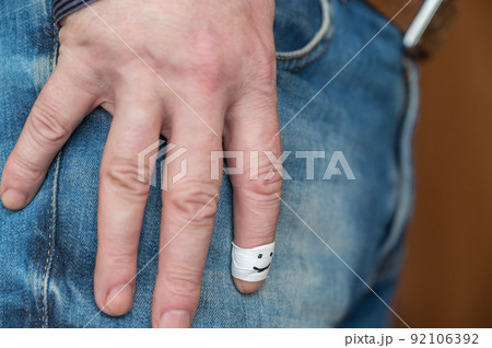 Male hand rests on his thigh. Middle-aged man is wearing blue jeans with brown leather belt. Index finger is wrapped in white tape. Happy smiling face is drawn in black marker on the finger. Male hand rests on his thigh. Middle-aged man is wearing blue jeans with brown leather belt. Index finger is wrapped in white tape. Happy smiling face is drawn in black marker on the finger. 92106392