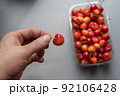 A man's hand holds a ripe sweet cherry berry.  A new crop of red berries in a transparent tray in the background. Healthy Food. Selective focus. 92106428