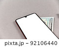 Smartphone and money on a gray screen. Smartphone with a white blank screen. A hundred-dollar bill lies under the phone. Copy space for text and design elements. Selective focus. 92106440