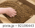 A woman hand picks up dry cat food. The food is in bulk in an open cardboard box. Triangular brown pellets. Pet supplies. Selective Focus. 92106443