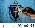 Witch flying on broomstick in front of Commonwealth of Massachusetts flag. Man hand holding figurine cut out of paper. Celebrating All Saints Night in United States concept. Selective focus. 92106444