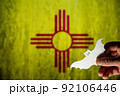 Happy Halloween, New Mexico. Male hand holds bat figurine. New Mexico state flag in background. Cut out of white paper Halloween paraphernalia. All Hallows evening. Selective focus. 92106446