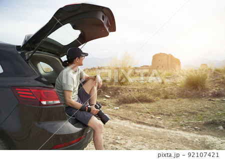 asian photographer sitting at the edge of trunk of a car looking at a deserted castle with camera in hand 92107421