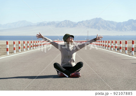 asian woman tourist sitting in the middle of an empty open road with lake and rolling mountains in background, leg crossed arms outstretched 92107885