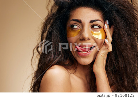 Enjoyed happy pretty Latin female with gold hydrogel patches under eye, stick tongue, looks up posing isolated over pastel beige background. Cosmetic product ad Natural beauty concept Studio portrait Enjoyed happy pretty Latin female with gold hydrogel patches under eye, stick tongue, looks up posing isolated over pastel beige background. Cosmetic product ad Natural beauty concept Studio portrait 92108125