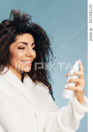 Smiling curly Latin lady in bathrobe look at toner spray in hand posing isolated on pastel blue background, feeling excited of effective skincare routine. Natural cosmetic ad concept. Copy space Smiling curly Latin lady in bathrobe look at toner spray in hand posing isolated on pastel blue background, feeling excited of effective skincare routine. Natural cosmetic ad concept. Copy space 92108126