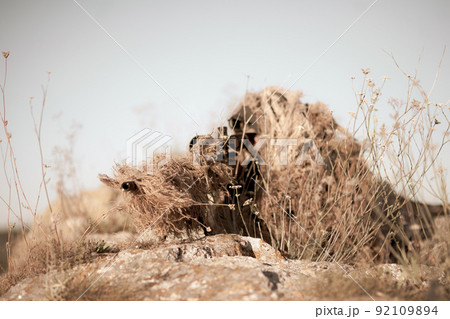 Special Forces Sniper in ghillie suit with a high-precision rifle with optic scope at combat position on the top of the rocky desert hillside Special Forces Sniper in ghillie suit with a high-precision rifle with optic scope at combat position on the top of the rocky desert hillside 92109894
